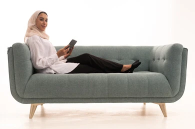 Using electronic programs and applications, having a good time on the mobile with friends, browsing social media sites, a portrait of a Saudi Arabian Gulf woman wearing a hijab in casual attire sitting on the couch sending text messages via mobile phone, white background.
