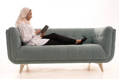 Browsing social media, having a good time on the phone with friends, a portrait of a Saudi Arabian Gulf woman wearing a hijab in casual attire sitting on the couch sending text messages via her mobile phone, white background.