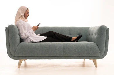 Spending enjoyable time on the mobile with friends, browsing social media, a portrait of a Saudi Arabian Gulf woman wearing a hijab and casual attire sitting on the couch sending text messages via her mobile phone, white background.