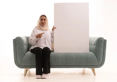 Marketing offers and advertisements, explanatory boards and educational tools, a portrait of a young Arab Gulf Saudi woman wearing a hijab in casual attire sitting on a sofa holding a blank rectangular white board with both hands and pointing at it, looking at the camera with expressions of happiness and joy, white rectangular mockup, white background.