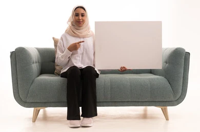 وسائط تعليمية ولوحات توضيحية، العروض والإعلانات التسويقية, بورتريه لشابة عربية خليجية سعودية محجبة ترتدي الزي الكاجوال تجلس على الاريكة تمسك بيديها لوحة بيضاء مستطيلة فارغة و تشير الية، النظر إلى الكاميرا بتعابير السعادة والسرور، موك اب مستطيل أبيض, خلفية بيضاء