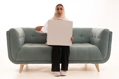Educational tools and explanatory boards, marketing presentations and advertisements, a portrait of a young Arab Gulf Saudi woman wearing a hijab in casual attire sitting on a sofa holding a blank rectangular white board with both hands and pointing at it, looking at the camera with expressions of happiness and joy, white rectangular mockup, white background.