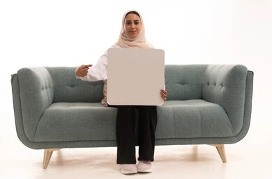 النظر إلى الكاميرا بتعابير السعادة والسرور، العروض والإعلانات التسويقية، لوحات توضيحية ووسائل تعليمية, بورتريه لشابة عربية خليجية سعودية محجبة ترتدي الزي الكاجوال تجلس على الاريكة تمسك بيديها لوحة بيضاء مستطيلة فارغة و تشير الية، موك اب مستطيل أبيض, خلفية بيضاء