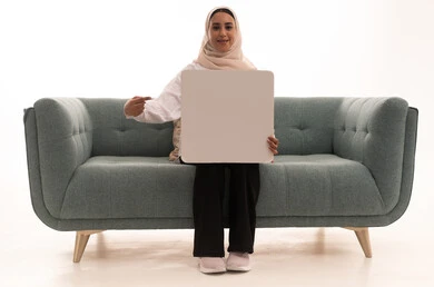 Illustrative boards and educational tools, marketing presentations and advertisements, a portrait of a young Arab Gulf Saudi woman wearing a hijab in casual attire sitting on a sofa holding a blank rectangular white board with both hands and pointing at it, looking at the camera with expressions of happiness and joy, white rectangular mockup, white background.