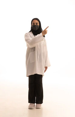 تحديد موقع معين, الدلالة والتوضيح على مكان عنصر ما, الالتزام بالاحترازات الوقائيه ضد كوفيد 19، بورتريه لطبيبة عربية خليجية سعودية ترتدي المعطف الطبي الأبيض وكمامة الوقاية تقوم بالإشارة إلى شيء محدد, الجسم بالطول الكامل, خلفية بيضاء