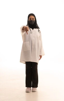 Discomfort about something, adherence to preventive measures against COVID-19, a portrait of an Arab Gulf Saudi female doctor wearing a medical coat, stethoscope, and mask, pointing her thumb down in gestures of disapproval and rejection, full-body, white background.