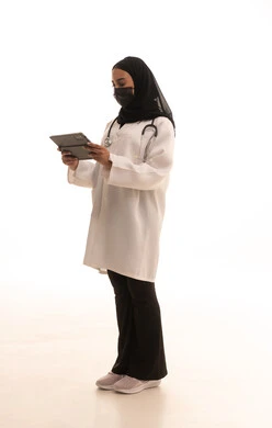 الحفاظ على السلامة من كوفيد19، تصفح مواقع الانترنت، الطب والرعاية الصحية، العمل في المراكز والمستشفيات، بورتريه لطبيبة عربية خليجية سعودية محجبة ترتدي المعطف الطبي الأبيض تضع سماعة الفحص الطبية و الكمامة تحمل التابلت، صورة لكامل الجسم، خلفية بيضاء