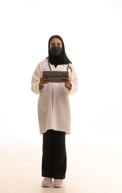 Browsing websites, medicine and healthcare, maintaining safety from COVID-19, working in centers and hospitals, a portrait of a veiled Arab Gulf Saudi female doctor wearing a white medical coat, holding a stethoscope and a mask, carrying a tablet, full-body image, white background.
