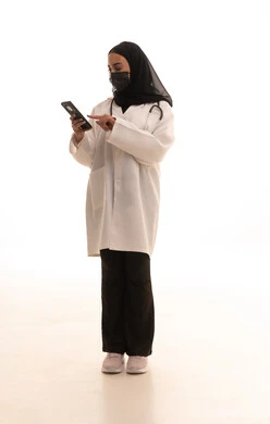 اتخاذ إجراءات السلامة للوقاية من كوفيد-19، التطبيقات الرقمية للخدمات الصحية، تقديم الخدمات والرعاية الصحية عبر الإنترنت، بورتريه لطبيبة عربيه خليجيه سعودية ترتدي المعطف الطبي و السماعه و الكمامة تقوم باستخدام الهاتف المحمول،الجسم بالطول الكامل، خلفية بيضاء