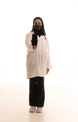 Maintaining safety from COVID-19, displaying a blank black screen, working in centers and hospitals, medicine and healthcare, a portrait of a Saudi Gulf Arab female doctor wearing a white medical coat and mask holding a mobile phone in her hand, a young Saudi woman providing medical assistance, full-body image, white background.