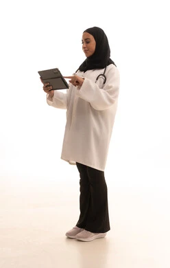 Providing remote consultations for patients using technological devices, the development of the healthcare sector in Saudi Arabia, working in the healthcare sector, a portrait of an Arab Gulf Saudi female doctor wearing a white coat and stethoscope holding a tablet device, full-body, white background.