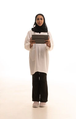 العمل في القطاع الصحي , تطور القطاع الصحي في المملكة السعودية،تقديم الاستشارات للمرضى عن بعد الأجهزة التكنولوجية، بورتريه لطبيبة عربية خليجية سعودية ترتدي المعطف الأبيض و السماعة تمسك جهاز التابلت ، الجسم بالطول الكامل, خلفية بيضاء