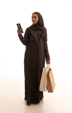 Browsing the internet via mobile phone, the positive effect of shopping on women's psychology, benefiting from seasonal offers and discounts, buying gifts and necessities, a portrait of a Saudi Gulf Arab woman wearing an abaya and hijab holding colorful bags in her hands with gestures of joy, full-body, white background.