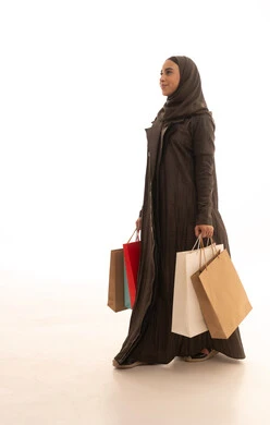 The positive effect of shopping on women's psychology, benefiting from seasonal offers and discounts, buying gifts and necessities, a side portrait of a Saudi Gulf Arab woman wearing an abaya and hijab holding colorful bags in her hands with gestures of joy, full-body, white background.