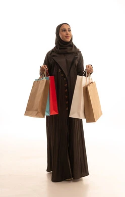 Looking at something, the positive effect of shopping on women's psychology, benefiting from seasonal offers and discounts, buying gifts and necessities, a portrait of a Saudi Gulf Arab woman wearing an abaya and hijab holding colorful bags in her hands with gestures of joy, full-body, white background.
