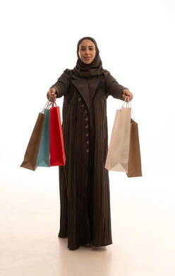 الاستفادة من العروض والتخفيضات الموسميه، شراء الهدايا والمستلزمات، الاُثر الايجابي للتسوق على نفسية النساء، بورتريه لامرأة عربية خليجية سعودية ترتدي العباءة والحجاب تحمل بيديها أكياس ملونة بإيماءات السرور, الجسم بالطول الكامل، خلفية بيضاء