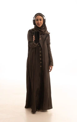 النظر الى الكاميرا بايماءات السعادة والسرور، بورتريه لإمرأة عربية خليجية سعودية مبتسمة ترتدي العباءة و الحجاب تضع الهيدفون تقوم بعرض شاشة سوداء فارغة عبر الهاتف المحمول،استخدام الأجهزة والتقنيات الحديثة والمتطورة، الجسم بالطول الكامل، خلفية بيضاء