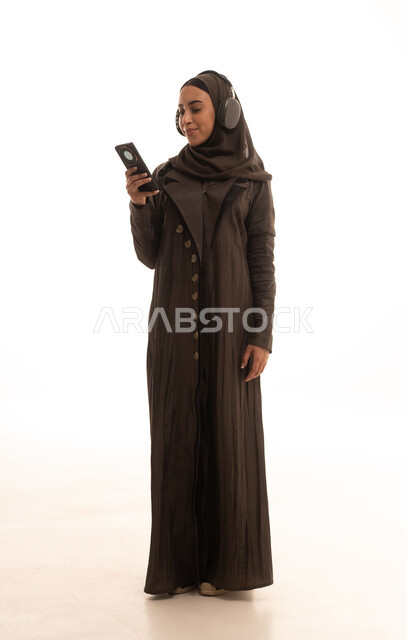 The use of advanced modern technologies, enjoying listening to favorite music, a portrait of a smiling Arab Saudi Gulf woman wearing a hijab, dressed in an abaya and headphones, using a mobile phone with gestures of joy, full-body, white background.