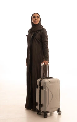 النظر الى شيء ما، الاستعداد و تجهيز الأمتعة للسفر في جولة سياحية، مفهوم السياحة و السفر، بورتريه من الجانب لامراه عربية خليجيه سعوديه مبتسمه تمسك حقيبة سفر، ايماءات وجه تدل على الحماس للقيام برحلة سياحية، الجسم بالطول الكامل، خلفية بيضاء