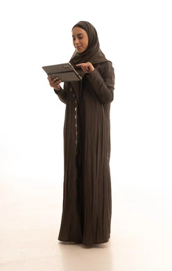 The concept of remote project completion, browsing websites and social media, integrating technology into daily and professional life, a portrait of a Saudi Gulf Arab woman wearing an abaya and hijab using a tablet, full-body, white background.