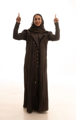 A specific location is identified, with hand signals and movements indicating this place (here), a portrait of a Saudi Arabian Gulf woman looking at the camera, raising her index fingers pointing upwards, full body, white background.