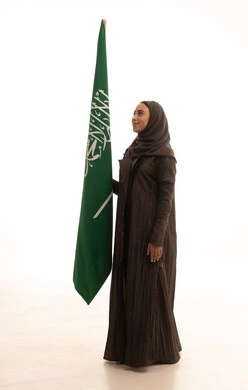 ذكرى يوم بدينا 1727 م، يوم العلم السعودي11 مارس، الاحتفال بتأسيس الدولة السعودية الأولى 22 فبراير، مفهوم حب الوطن و الانتماء اليه، بورتريه من الجانب لإمرأة عربية خليجية سعودية ترتدي العباءة و الحجاب تمسك راية وشعار المملكة تنظر بايماءات الاعتزاز و الفخر, الجسم بالطول الكامل, خلفية بيضاء