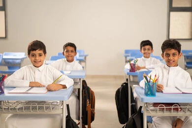 Performing school assignments, preparing for the new school year, education in Saudi Arabia, back to school, a group of Saudi Gulf Arab students wearing traditional attire sitting at their desks and doing their homework, a Saudi Gulf Arab teacher wearing traditional attire and a shemagh standing next to his students.