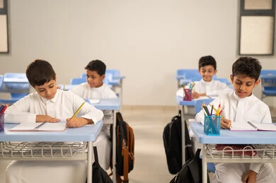 Performing school assignments, preparing for the new school year, education in Saudi Arabia, back to school, a group of Saudi Gulf Arab students wearing traditional attire sitting at their desks and doing their homework, a Saudi Gulf Arab teacher wearing traditional attire and a shemagh standing next to his students.