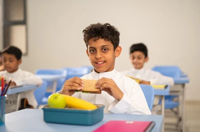 مجموعة من الطلاب العربيين الخليجيين السعوديين يجلسون في مقاعدهم وأمامهم اللانش البوكس، الوجبات الغنية بالعناصر الغذائية الهامة لبناء الجسمK الاستمتاع بتناول الأطعمة الصحية في وقت الفسحة المدرسية، موسم العودة للمدارس