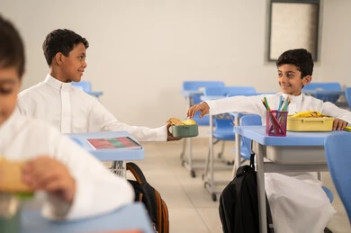 Sharing food with gestures of affection and familiarity, expressions of happiness and joy, enjoying healthy foods during school break time, a group of Arab Gulf Saudi students sitting in their seats with lunch boxes in front of them, meals rich in essential nutrients for building the body.