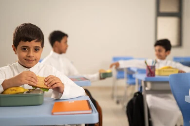 Sharing food with gestures of affection and familiarity, expressions of happiness and joy, enjoying healthy foods during school break time, a group of Arab Gulf Saudi students sitting in their seats with lunch boxes in front of them, meals rich in essential nutrients for building the body.