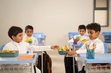 Sharing food with gestures of affection and familiarity, expressions of happiness and joy, enjoying healthy foods during school break time, a group of Arab Gulf Saudi students sitting in their seats with lunch boxes in front of them, meals rich in essential nutrients for building the body.