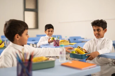 Sharing food with gestures of affection and familiarity, expressions of happiness and joy, enjoying healthy foods during school break time, a group of Arab Gulf Saudi students sitting in their seats with lunch boxes in front of them, meals rich in essential nutrients for building the body.