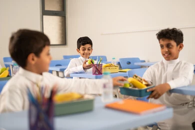 مشاركة الطعام بايماءات الود والالفه، تعابير السعاده والسرور ، الاستمتاع بتناول الأطعمة الصحية في وقت الفسحة المدرسية، مجموعة من الطلاب العربيين الخليجيين السعوديين يجلسون في مقاعدهم وأمامهم اللانش البوكس، الوجبات الغنية بالعناصر الغذائية الهامة لبناء الجسم