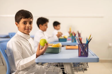 الوجبات الغنية بالعناصر الغذائية الهامة لبناء الجسم، الاستمتاع بتناول الأطعمة الصحية في وقت الفسحة المدرسية، موسم العودة للمدارس، مجموعة من الطلاب العربيين الخليجيين السعوديين يجلسون في مقاعدهم وأمامهم اللانش البوكس