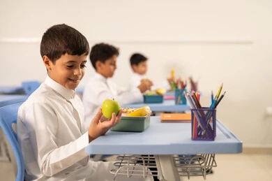 موسم العودة للمدارس، الاستمتاع بتناول الأطعمة الصحية في وقت الفسحة المدرسية،مجموعة من الطلاب العربيين الخليجيين السعوديين يجلسون في مقاعدهم وأمامهم اللانش البوكس، الوجبات الغنية بالعناصر الغذائية الهامة لبناء الجسم