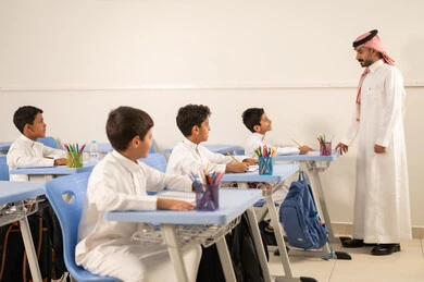The quality of education in Saudi Arabia, a Gulf Arab Saudi teacher wearing traditional attire and a ghutrah discussing lessons with his students, a group of Gulf Arab Saudi students in school uniforms doing their homework, active participation of students with teachers, education and teaching with the prescribed curricula for students, friendly dialogues between the teacher and students.
