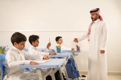 التربية والتعليم بالمناهج المقررة للطلاب في المملكة العربية السعودية، تعابير وايماءات الحماس لمعرفة الجواب الصحيح، طالب عربي خليجي سعودي مبتسم يرتدي الثوب التقليدي يرفع يديه للإجابة يمسك بيده قلم ، التفاعل والمشاركة داخل الفصل الدراسي