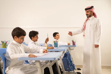 طالب عربي خليجي سعودي مبتسم يرتدي الثوب التقليدي يرفع يديه للإجابة يمسك بيده قلم ، التفاعل والمشاركة داخل الفصل الدراسي، التربية والتعليم بالمناهج المقررة للطلاب في المملكة العربية السعودية، تعابير وايماءات الحماس لمعرفة الجواب الصحيح