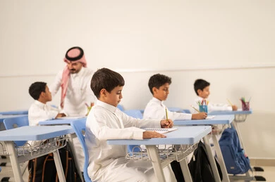 The quality of education in Saudi Arabia, a Gulf Arab Saudi teacher wearing traditional attire and a ghutrah discussing lessons with his students, a group of Gulf Arab Saudi students in school uniforms doing their homework, active participation of students with teachers, education and teaching with the prescribed curricula for students, friendly dialogues between the teacher and students.