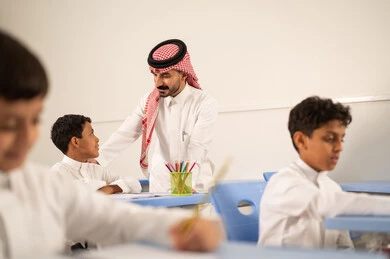 The quality of education in Saudi Arabia, a Gulf Arab Saudi teacher wearing traditional attire and a ghutrah discussing lessons with his students, a group of Gulf Arab Saudi students in school uniforms doing their homework, active participation of students with teachers, education and teaching with the prescribed curricula for students, friendly dialogues between the teacher and students.