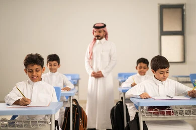 Performing school assignments, preparing for the new school year, education in Saudi Arabia, back to school, a group of Saudi Gulf Arab students wearing traditional attire sitting at their desks and doing their homework, a Saudi Gulf Arab teacher wearing traditional attire and a shemagh standing next to his students.