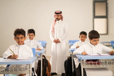 Performing school assignments, preparing for the new school year, education in Saudi Arabia, back to school, a group of Saudi Gulf Arab students wearing traditional attire sitting at their desks and doing their homework, a Saudi Gulf Arab teacher wearing traditional attire and a shemagh standing next to his students.