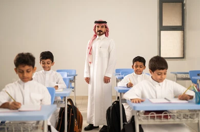 Performing school assignments, preparing for the new school year, education in Saudi Arabia, back to school, a group of Saudi Gulf Arab students wearing traditional attire sitting at their desks and doing their homework, a Saudi Gulf Arab teacher wearing traditional attire and a shemagh standing next to his students.