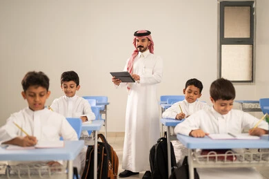 The use of modern technologies, completing school assignments, preparing for the new school year, returning to school, a group of Saudi Gulf Arab students wearing traditional attire sitting at their desks and doing their homework, a Saudi Gulf Arab teacher wearing traditional clothing and a ghutrah holding a tablet stands next to his students.