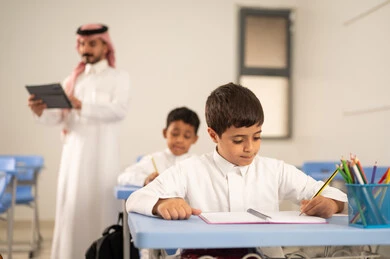 The use of modern technologies, completing school assignments, preparing for the new school year, returning to school, a group of Saudi Gulf Arab students wearing traditional attire sitting at their desks and doing their homework, a Saudi Gulf Arab teacher wearing traditional clothing and a ghutrah holding a tablet stands next to his students.