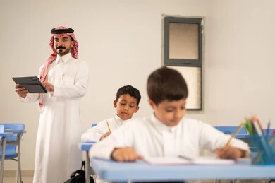 العودة إلى المدارس، استخدام التقنيات الحديثة، تأدية الواجبات المدرسية، الإستعداد للعام الدراسي الجديد، مجموعة من الطلاب العربيين الخليجيين السعوديين يرتدون الزي التقليدي  يجلسون في مقاعدهم ويقومون بحل الواجب ، معلم عربي خليجي سعودي يرتدي الزي التقليدي والشماغ يمسك بيده جهاز التابلت يقف بجانب طلابه