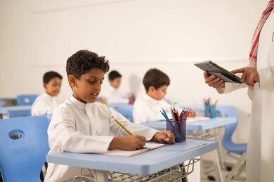 The use of modern technologies, a Saudi Gulf Arab teacher wearing traditional attire and a shemagh is discussing lessons with his students while holding a tablet in his hand. A group of Gulf Arab Saudi students wearing school uniforms are doing their homework, actively participating with the teachers, and having friendly dialogues between the teacher and the students.