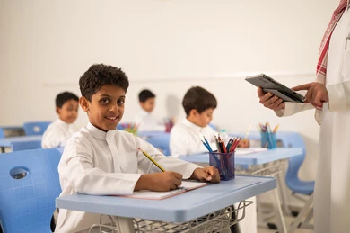 معلم عربي خليجي سعودي يرتدي الثوب التقليدي والشماغ يقوم بمناقشة الدروس مع طلابه يمسك بيده جهاز التابلت ، مجموعة من طلاب عربيين خليجيين سعوديين يرتدون الزي المدرسي يقومون بتأديه الواجبات ، المشاركة الفعالة للطلاب مع المدرسين،حوارات ودية بين الاستاذ والطلاب، استخدام التقنيات الحديثة