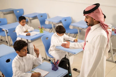Expressions and gestures of excitement to know the correct answer, a smiling Saudi Gulf Arab student wearing traditional attire raises his hands to answer, interaction and participation inside the classroom, education and teaching with the prescribed curricula for students in the Kingdom of Saudi Arabia.